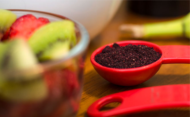 A bowl of assorted fruit next to a teaspoon measure filled with powdered wild blueberries.